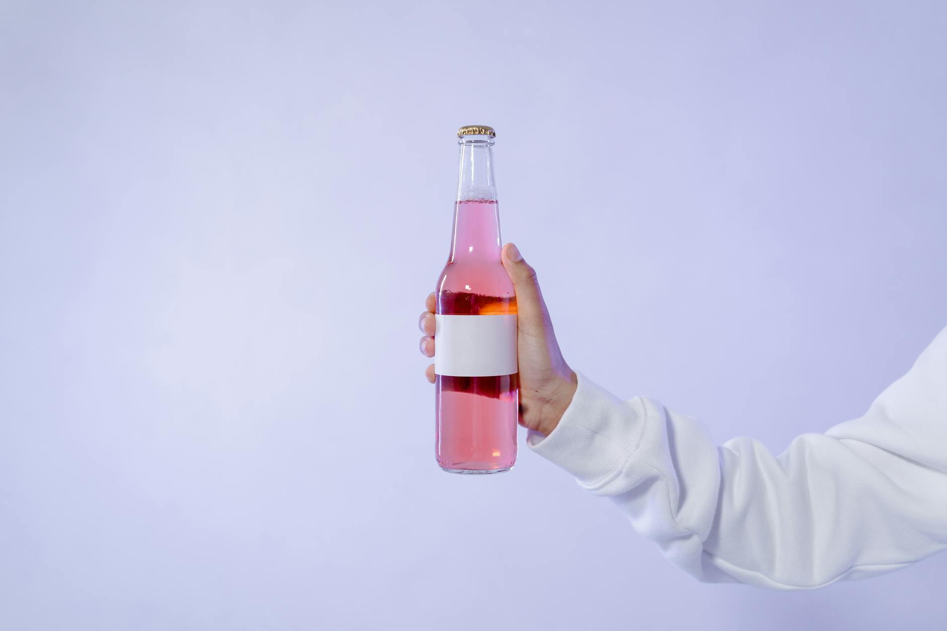 Botella de bebida rosa sostenida por una mano sobre fondo claro.